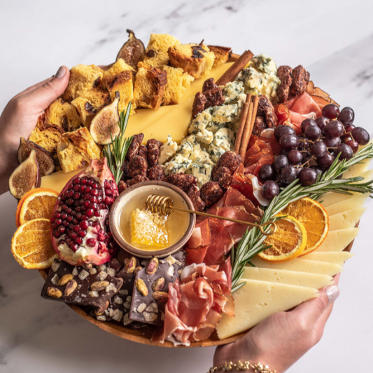 Platter of assorted meats, cheeses, fruits, and bread held by two hands on a marble surface.