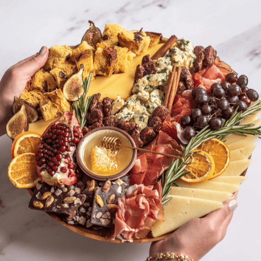 Platter of assorted meats, cheeses, fruits, and bread held by two hands on a marble surface.