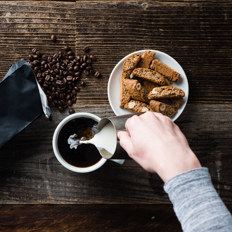 Person adding cream to a cup of coffee on a wooden table with coffee beans and cocoa biscotti.