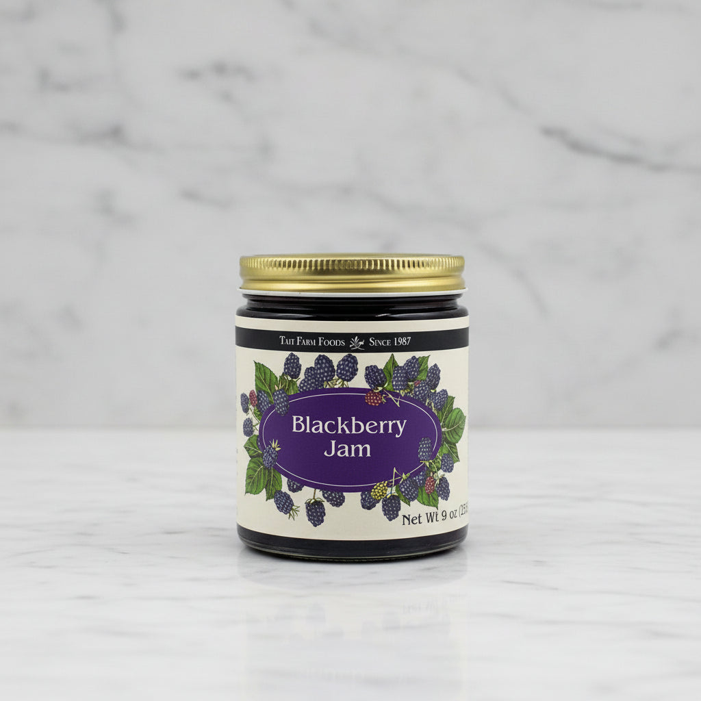 A jar of TaiT Farm Foods Blackberry Jam with a gold metal lid and a cream-colored label decorated with illustrations of blackberries and green leaves.