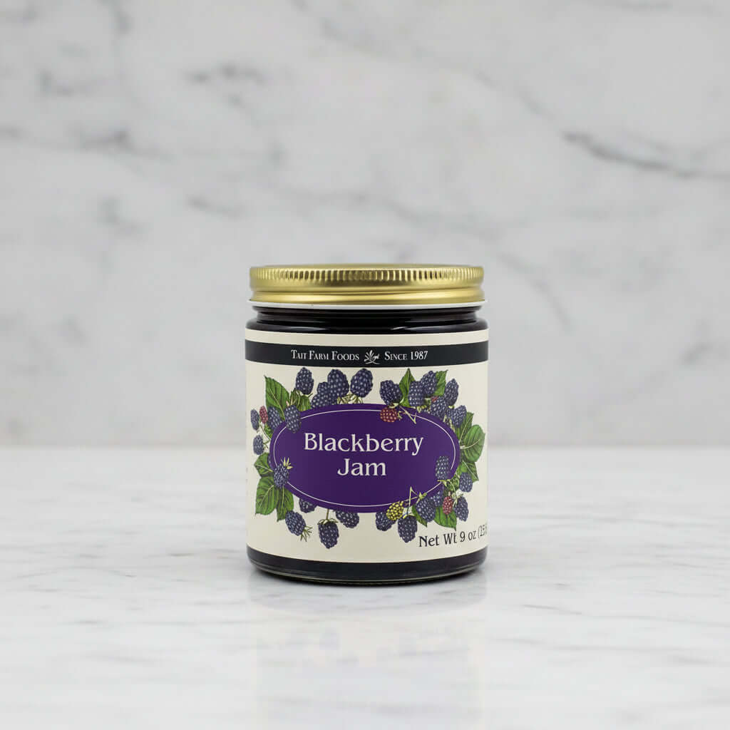 A jar of TaiT Farm Foods Blackberry Jam with a gold metal lid and a cream-colored label decorated with illustrations of blackberries and green leaves.