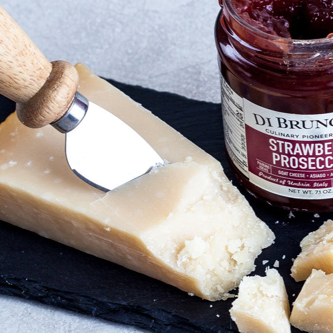 Jar of Di Bruno Bros Strawberry & Prosecco Jam with a block of cheese and a cheese knife on a slate board.