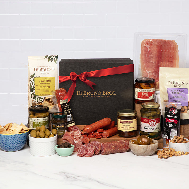 A gourmet gift box from Di Bruno Bros. containing an assortment of meats, cheeses, and crackers arranged neatly inside the box, tied with a red ribbon.
