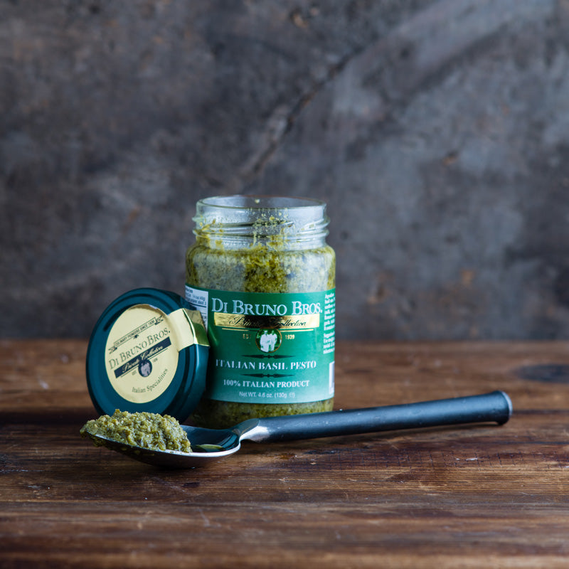 A jar of Di Bruno Bros. pesto sauce with its lid removed, alongside a spoon with pesto on it, all placed on a wooden surface.