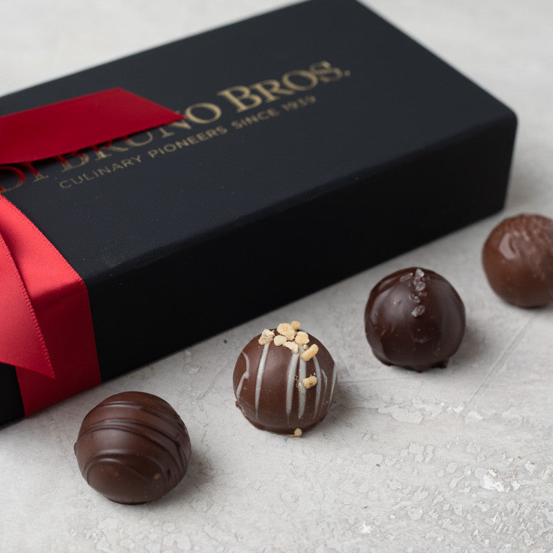 An assortment of Di Bruno Bros chocolate truffles with different toppings such as nuts and caramel, displayed in front of their black packaging with a red ribbon.