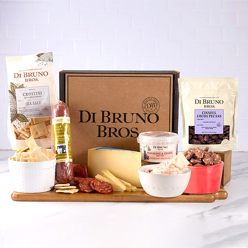 A gourmet gift box from Di Bruno Bros. containing a variety of cheeses, charcuterie, and candied nuts displayed on a wooden board, with packaging and additional items like crostini and salami.