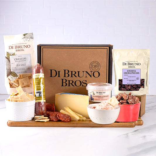 A gourmet gift box from Di Bruno Bros. containing a variety of cheeses, charcuterie, and candied nuts displayed on a wooden board, with packaging and additional items like crostini and salami.