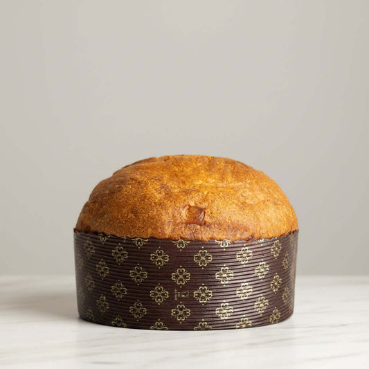 A round golden-brown panettone loaf in a dark brown paper liner with a repeating gold floral pattern, photographed against a plain light marble background.