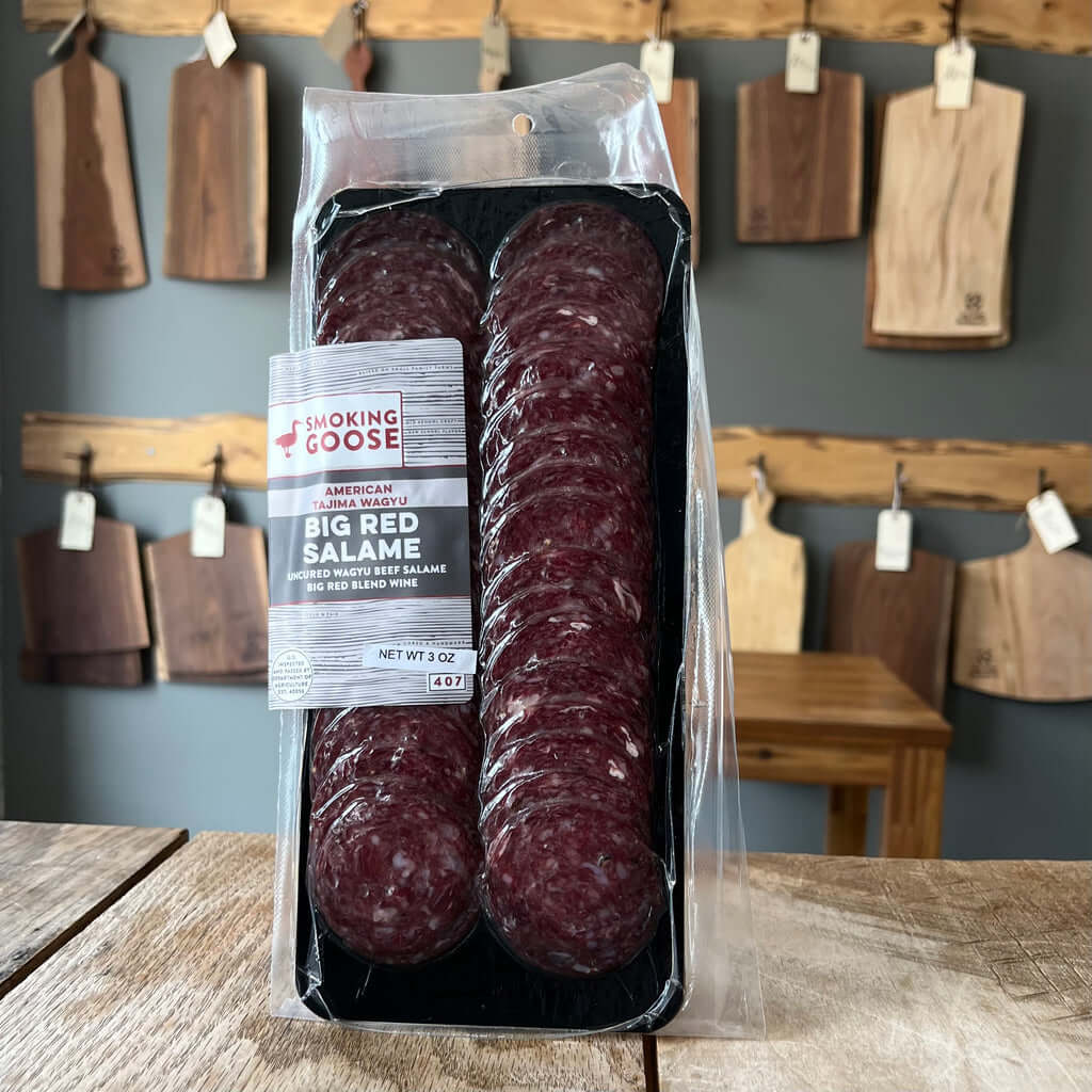 Big Red Wagyu Salame in clear packaging, sliced rounds on a wooden table, with rustic kitchen backdrop.