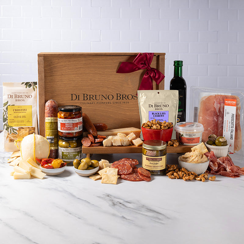 An assortment of gourmet food items from Di Bruno Bros. Italian Market-style pepperoni, artisan candied nuts, mini provolone ball, prosciutto-stuffed pepper shooters, roasted peppers, Cerignola olives, classico balsamic vinegar, salami, cheese spread, and crostini are displayed around a wooden gift crate with a red ribbon.