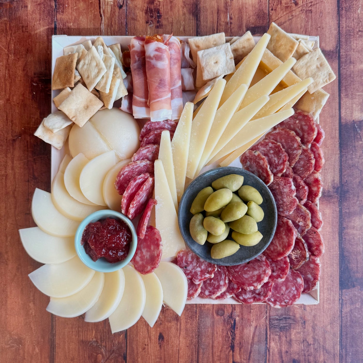 An antipasto platter featuring an assortment of Italian delicacies including prosciutto, soppressata, cheese slices, crostini, olives, and jam, arranged on a cheese board.