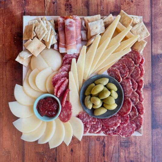 An antipasto platter featuring an assortment of Italian delicacies including prosciutto, soppressata, cheese slices, crostini, olives, and jam, arranged on a cheese board.