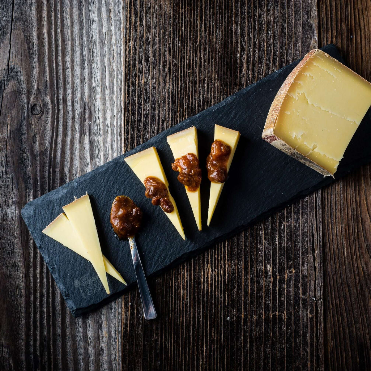 Cheese platter with sliced cheese and condiments on a dark slate board over a wooden background