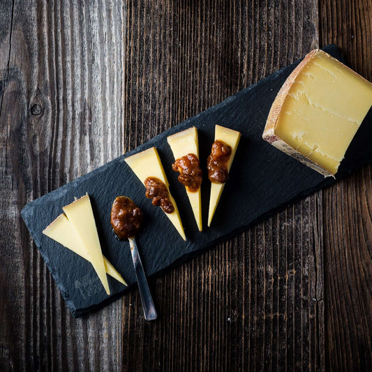 Cheese platter with sliced cheese and condiments on a dark slate board over a wooden background