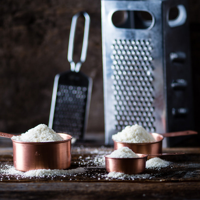 A photo showing grated Parmesan cheese in a copper bowl with a grater in the background, all placed on a wooden surface.