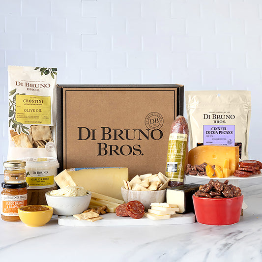 An assortment of gourmet cheese, charcuterie, and accessories presented in a gift box with Di Bruno Bros. branding. Items include cheese spreads, aged cheddar, piave cheese, candied nuts, jam, gouda cheese, crostini, and white truffle honey.