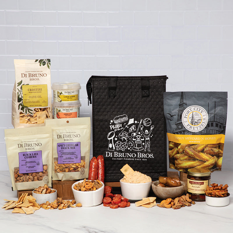 A Tailgate bundle of gourmet snacks from Di Bruno Bros including pepper shooters, cheese spreads, candied nuts, snack mix, crackers, pretzel sticks, and pepperoni, displayed on a table with a black Di Bruno Bros. bag.