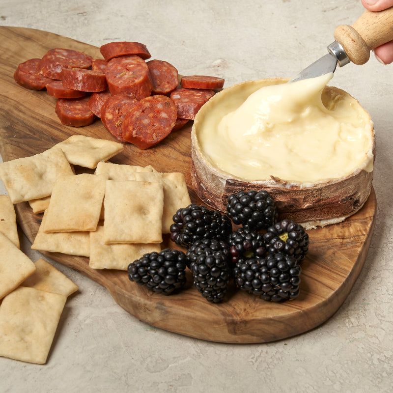 A wheel of Harbison cheese with a white rind, partially unwrapped from its spruce bark casing, sitting on a wooden board with a cheese knife.