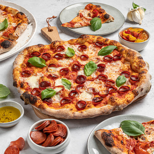 A pizza kit from Di Bruno Bros. with various ingredients displayed, including dough balls, crushed tomatoes, mozzarella cheese, roasted peppers, pepperoni, and parmesan flakes, along with a prepared pizza topped with cheese, pepperoni, and sauce.