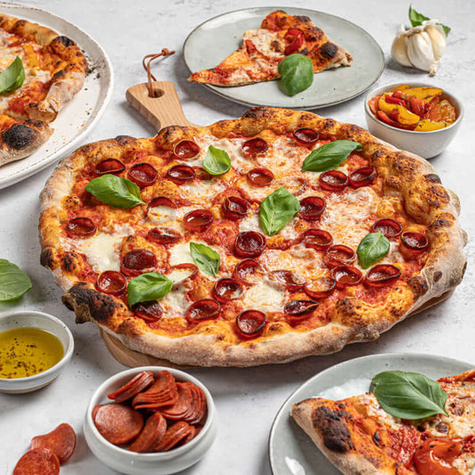 A pizza kit from Di Bruno Bros. with various ingredients displayed, including dough balls, crushed tomatoes, mozzarella cheese, roasted peppers, pepperoni, and parmesan flakes, along with a prepared pizza topped with cheese, pepperoni, and sauce.