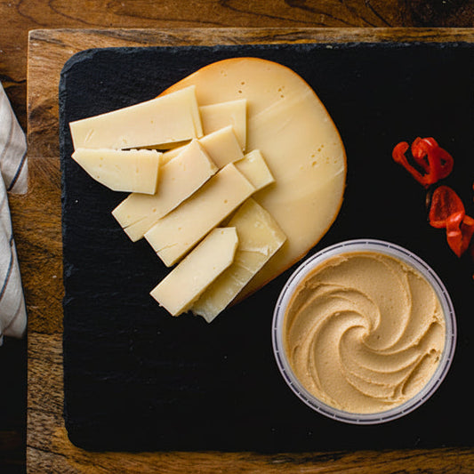 Cheese platter with sliced cheese, a container of cheese spread, and red peppers on a dark slate board.