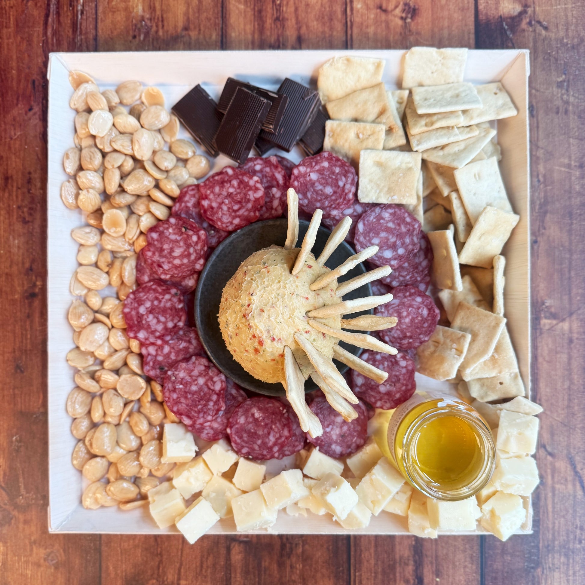 An overhead photo of a charcuterie board featuring an assortment of gourmet treats including sliced salami, cheese spread, crackers, almonds, and honey with a cheese board and knife set.