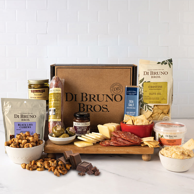 An assortment of gourmet cheese, charcuterie, and accoutrements displayed around a cardboard gift box with 'Di Bruno Bros' branding.