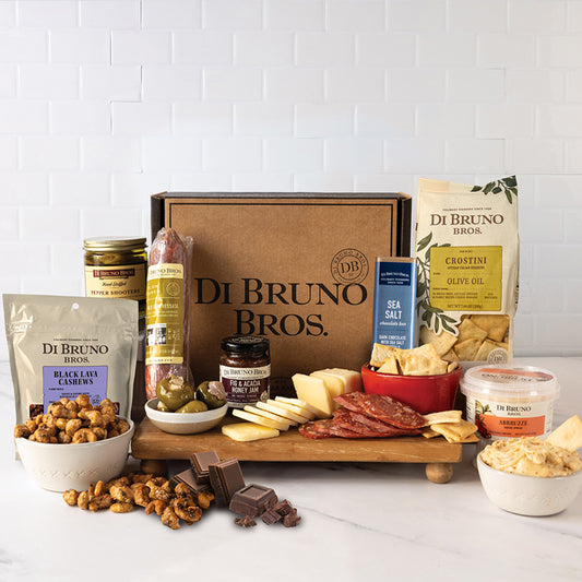 An assortment of gourmet cheese, charcuterie, and accoutrements displayed around a cardboard gift box with 'Di Bruno Bros' branding.
