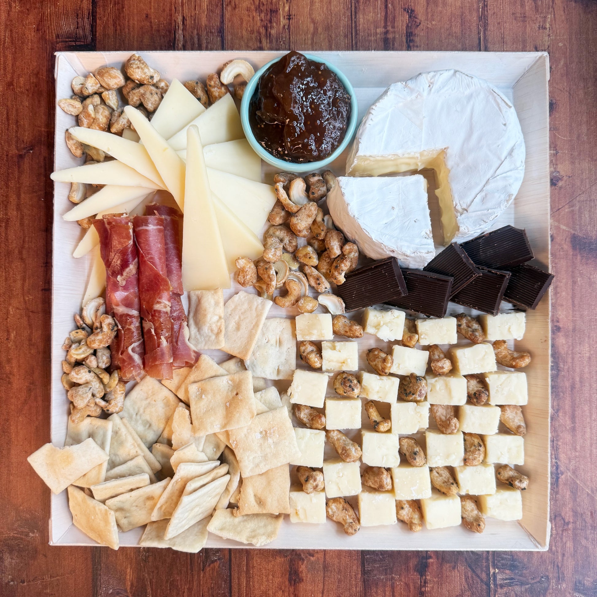 An overhead photo of an assembled charcuterie board featuring an array of cheeses, meats, nuts, crackers, and a small bowl of jam, arranged neatly on a cheese board.