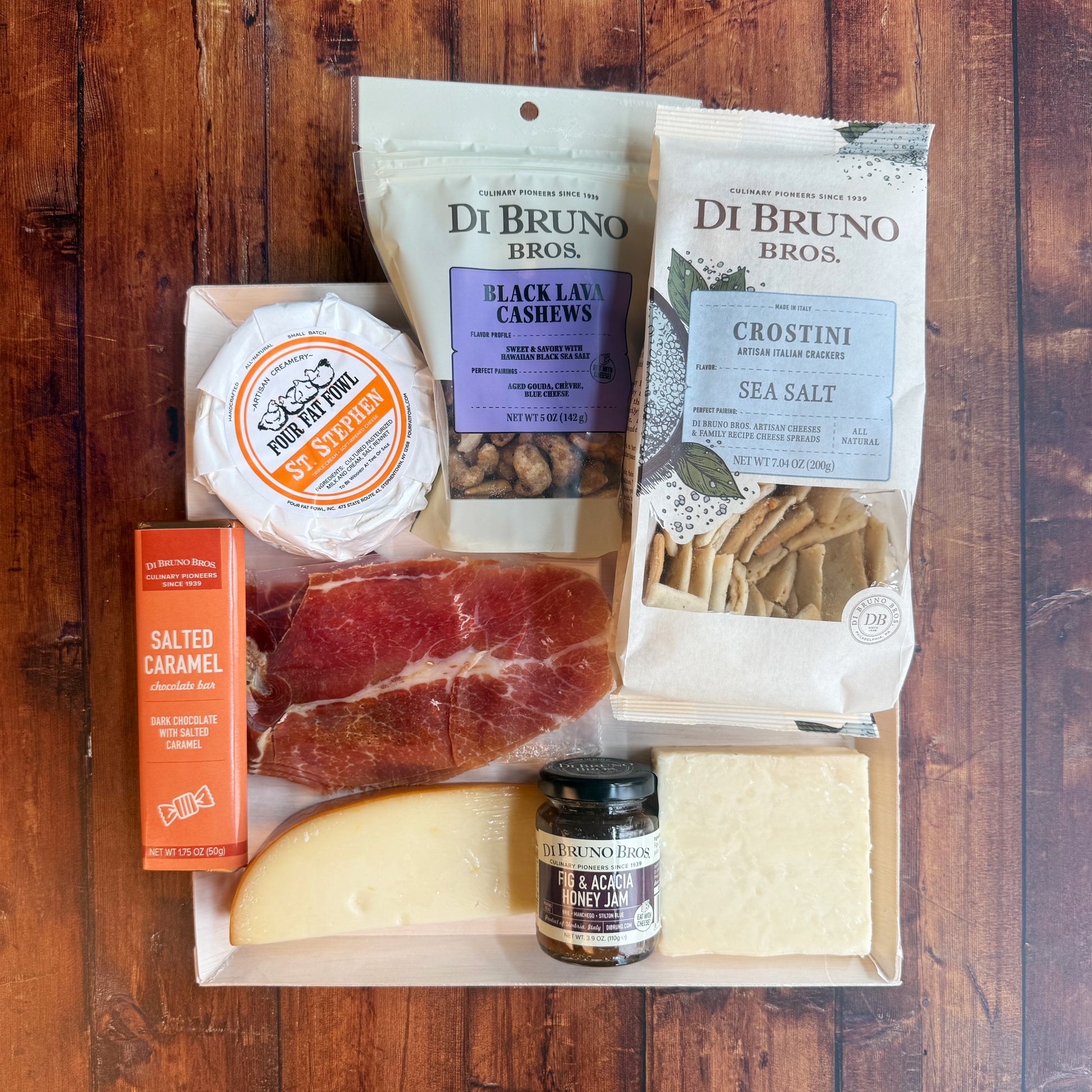 An overhead photo of a packaged charcuterie board featuring an array of cheeses, meats, nuts, crackers, and a small bowl of jam, arranged placed on a board.