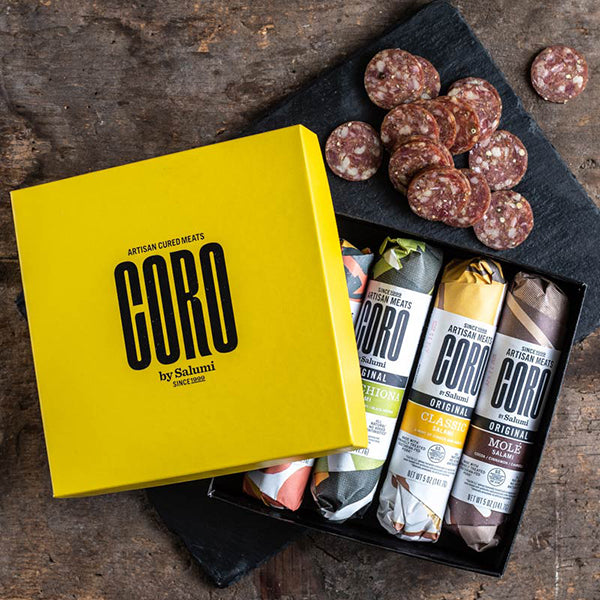 A gift box of Coro by Salumi salamis, with an assortment of sliced salami visible next to the box on a wooden surface.