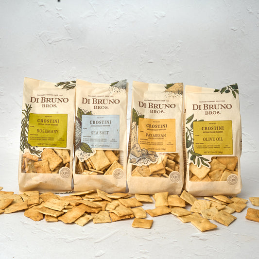 Four packages of Di Bruno Bros. Crostini Sampler Set with an assortment of flavors including olive oil, parmesan, rosemary, and sea salt, displayed in front of a white background with scattered crostini crackers.