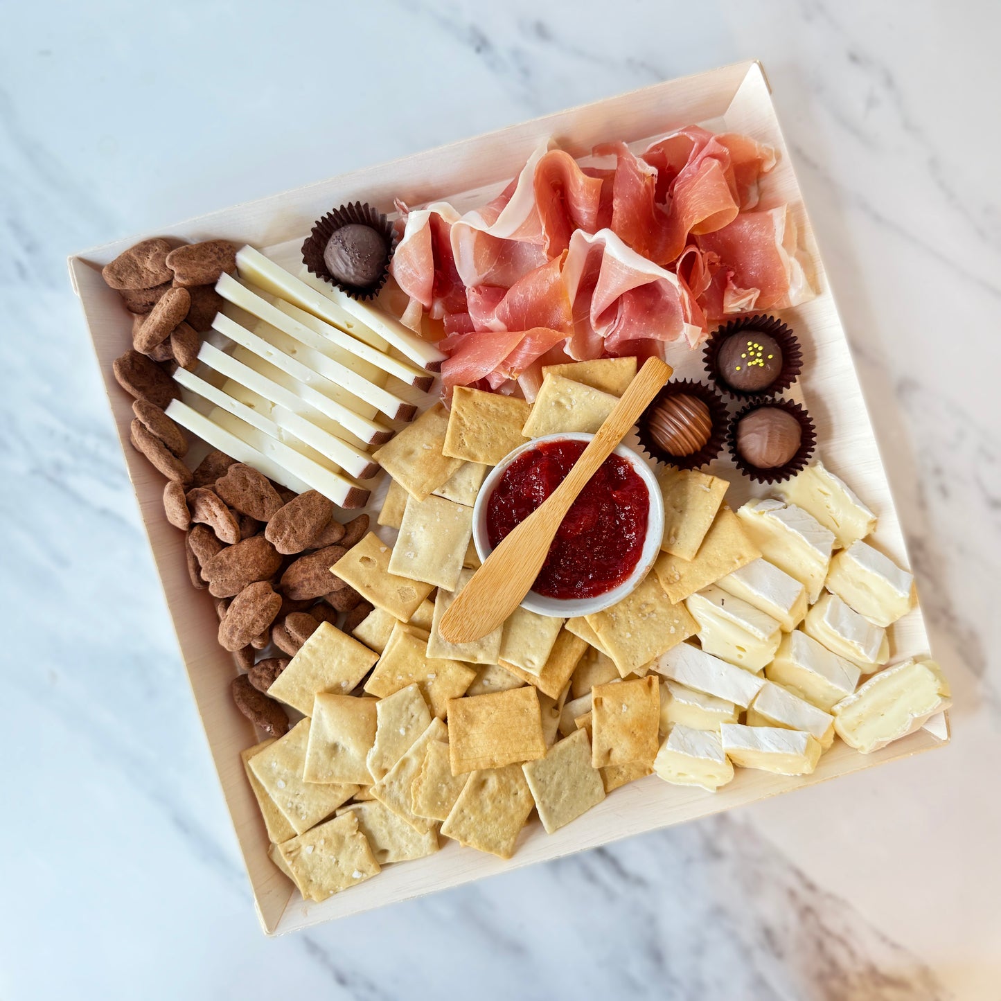Assorted charcuterie board with meats, cheeses, crackers, and chocolates on a marble surface.