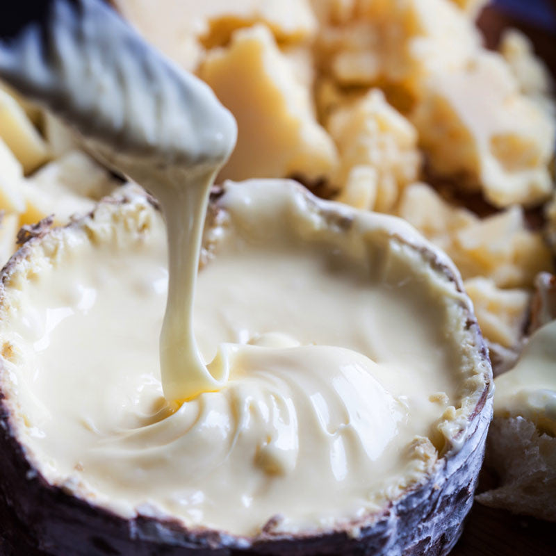 A wheel of Harbison cheese with a white rind, partially unwrapped from its spruce bark casing, sitting on a wooden board with a cheese knife.