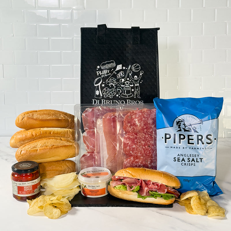 An Italian Hoagie Kit displayed with hoagie rolls, prosciutto, coppa, salame, roasted peppers, cheese spread, and potato chips.