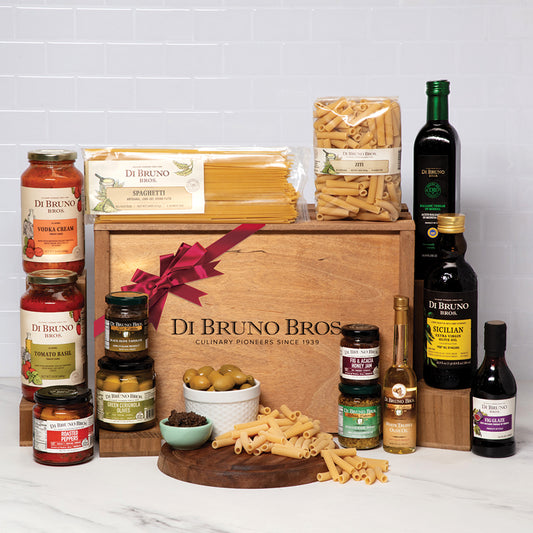A gourmet gift crate from Di Bruno Bros. featuring an assortment of pasta, sauces, olive oils, antipasti, and other food items, presented in a wooden crate with a red bow.