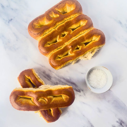 Two pretzel-shaped bread loaves on a marble surface with a small bowl of salt.