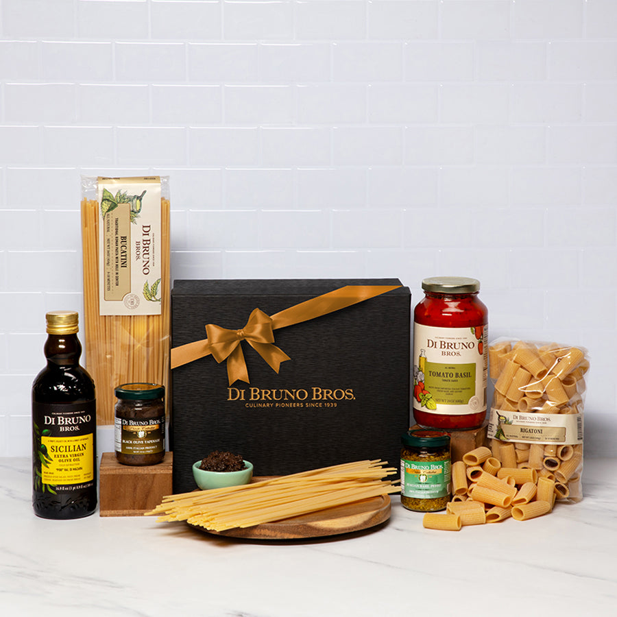 A gourmet gift box from Di Bruno Bros. containing various pasta-related items including olive oil, rigatoni pasta, black olive tapenade, tomato basil pasta sauce, bucaturi pasta, and pesto sauce, all displayed neatly on a white background.