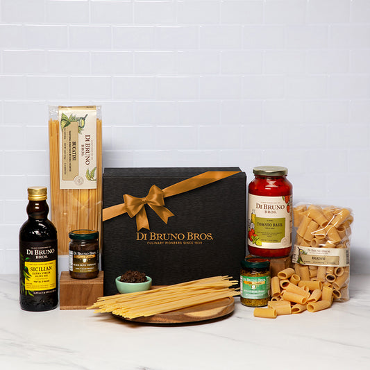 A gourmet gift box from Di Bruno Bros. containing various pasta-related items including olive oil, rigatoni pasta, black olive tapenade, tomato basil pasta sauce, bucaturi pasta, and pesto sauce, all displayed neatly on a white background.