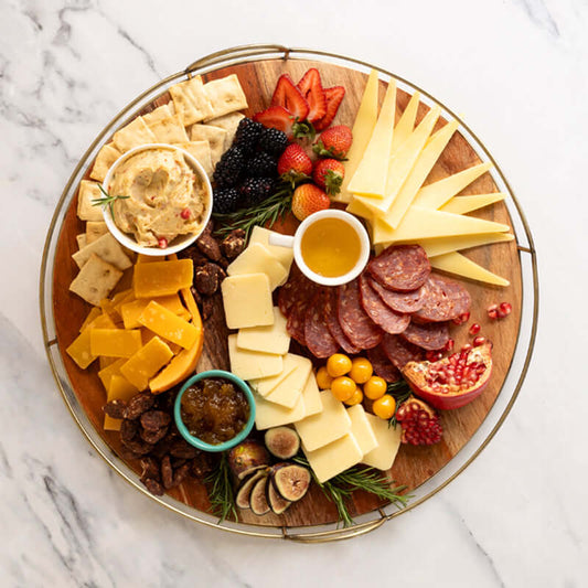 An assortment of gourmet cheese, charcuterie, and accessories presented in a gift box with Di Bruno Bros.. Items include cheese spreads, aged cheddar, piave cheese, candied nuts, jam, gouda cheese, crostini, and white truffle honey.