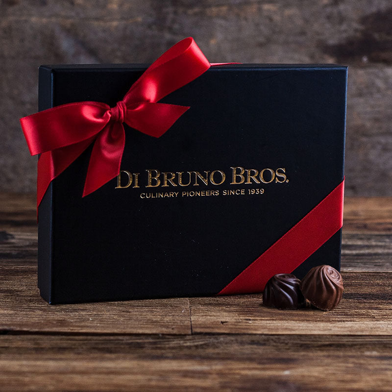 A box of Di Bruno Bros. chocolate truffles with a red ribbon on a wooden surface, with two truffles in front of the box.