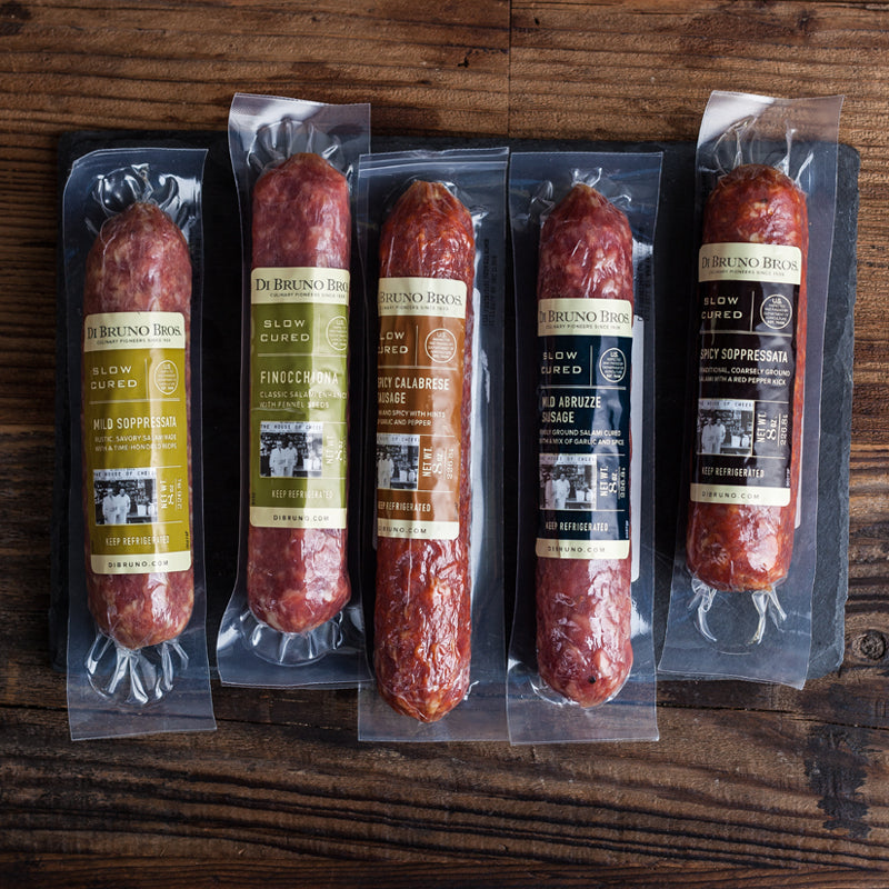 A set of packaged salami varieties from Di Bruno Bros., including Italian Market-Style Pepperoni, Sicilian Pepperoni, Mild Soppressata, Spicy Soppressata, and Finocchiona, displayed on a wooden surface.