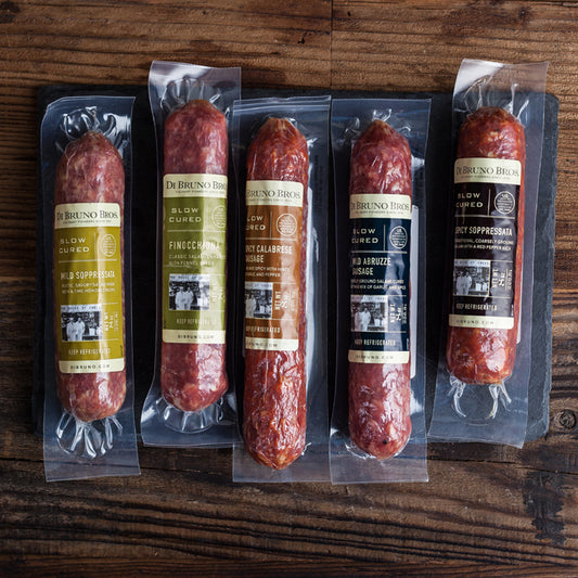A set of packaged salami varieties from Di Bruno Bros., including Italian Market-Style Pepperoni, Sicilian Pepperoni, Mild Soppressata, Spicy Soppressata, and Finocchiona, displayed on a wooden surface.