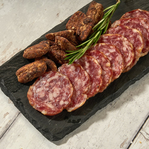 A slate serving board with slices of sweet soppressata salami and what appears to be prosciutto, garnished with a sprig of herbs.