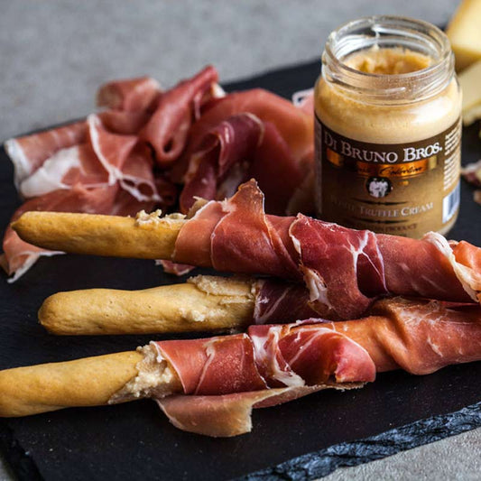 Prosciutto-wrapped bread sticks with a jar of truffle cream on a black board.