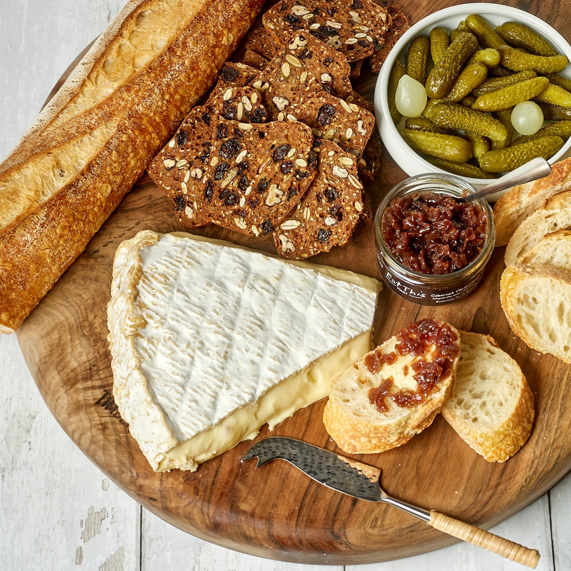 Wooden cutting board with bread, cheese, and pickles on a light wooden surface