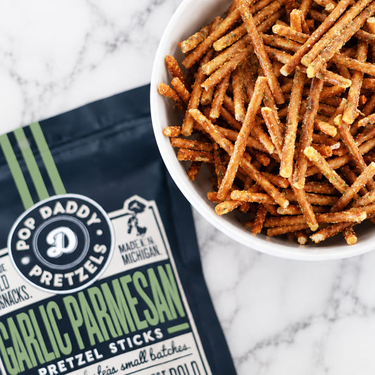 A white bowl filled with Pop Daddy Pretzel Sticks garlic parmesan snack sticks, shown next to the dark navy and green branded bag on a patterned cloth over a wooden surface.