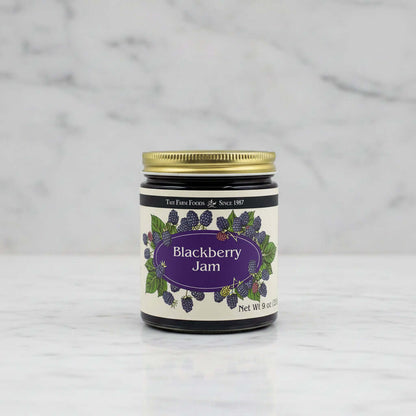 A jar of TaiT Farm Foods Blackberry Jam with a gold metal lid and a cream-colored label decorated with illustrations of blackberries and green leaves.