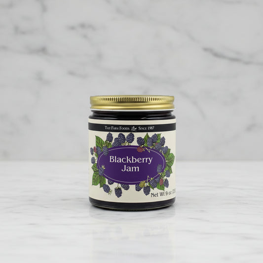 A jar of TaiT Farm Foods Blackberry Jam with a gold metal lid and a cream-colored label decorated with illustrations of blackberries and green leaves.