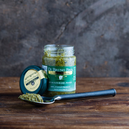 A jar of Di Bruno Bros.  pesto sauce with its lid removed, alongside a spoon with pesto on it, all placed on a wooden surface.
